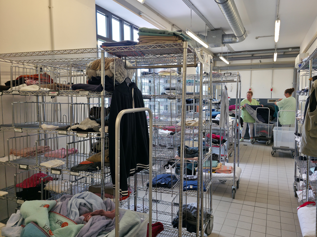 Unsere Wäscherei in der Totalen. Im Vordergrund stehen die Wäscheregale und im Hintergrund stehen zwei Mitarbeiterinnen an den Waschmaschinen und leeren diese.