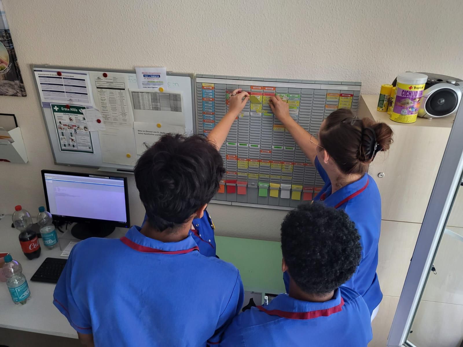 Drei Azubis, mit dem Rücken zum Bild, stehen an der Dienstplantafel.